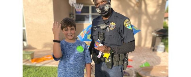 Police officer standing next to young male who is smiling and holding up his right arm to show bracelet that officer gave to him